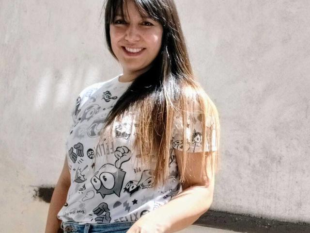 RoxanneBlair with long brown hair, smiling, wearing a white graphic tee with cartoon characters, standing against a plain wall, showing her small tits and slim waist.