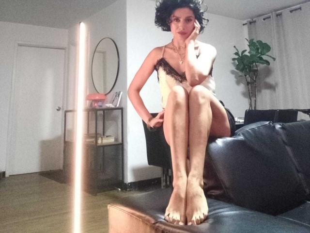 LilaaSalmon mit lockigem, schwarzem Haar, trägt ein zartes, gelb-schwarzes Dessous-Oberteil und sitzt auf einer schwarzen Ledercouch, die Beine gekreuzt, eine Hand am Kinn, im Hintergrund eine Pflanze..