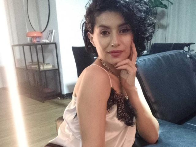 Lilaa, eine Frau mit kurzen, lockigen, schwarzen Haaren, trägt eine weiße, mit Spitze besetzte Trägertop und lehnt ihr Kinn in ihre Hand, lächelnd, während sie auf einem blauen Sofa sitzt..