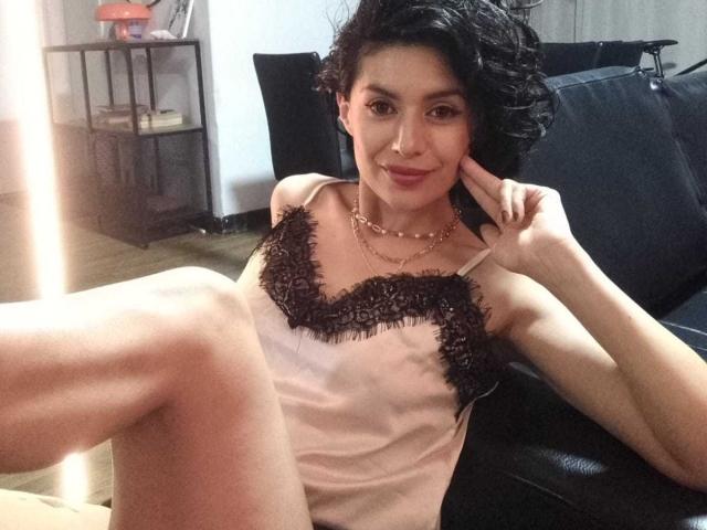 LilaaSalmon mit kurzen, schwarzen Haaren, trägt ein pinkes Tanktop mit Spitzenbesatz, sitzt auf einer schwarzen Couch, die Beine gespreizt, lächelt, eine Hand am Wangenknochen, trägt eine goldene Halskette..
