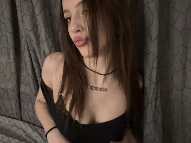 SimonneBrustkern with long brown hair, pink lips, black off-shoulder top showing small tits, tattoo on chest, black necklace, grey curtain background, looking slightly to the side.