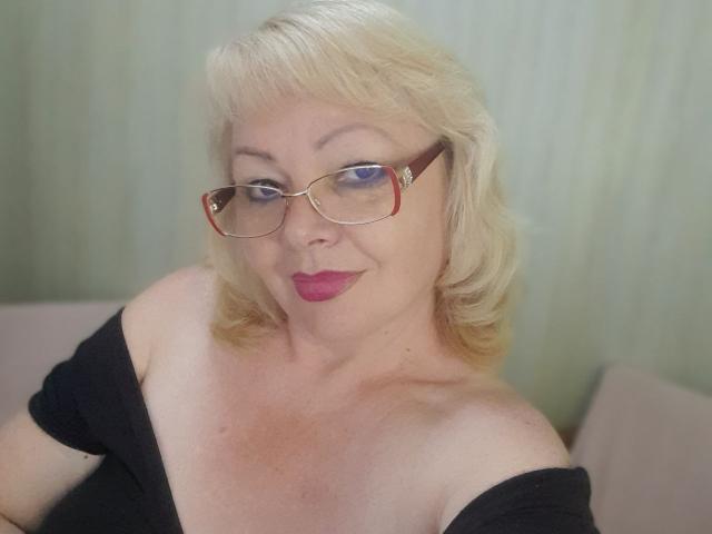Blonde EvelynLoveShy with glasses, red lipstick, and black off-shoulder top, showing her tits and smiling. She has fair skin and short, wavy hair.