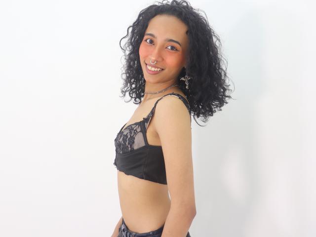 Agnis, avec ses cheveux noirs bouclés, sa peau bronzée et un anneau de nez, portant de la lingerie en dentelle noire, sourit à la caméra, montrant de petites seins et un joli visage..