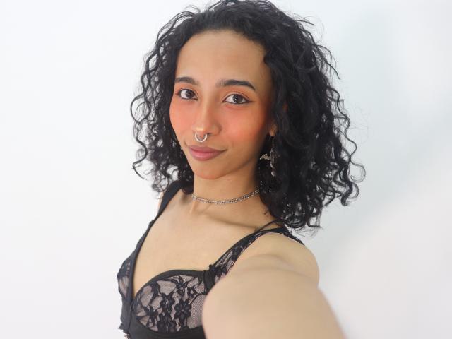 Agnis, avec ses cheveux noirs bouclés, sa peau brune, un anneau au nez et du rouge à lèvres rose, porte un soutien-gorge en dentelle noire et prend un selfie. Ses seins sont visibles, de taille moyenne. Elle porte un collier ras du cou. Elle sourit..