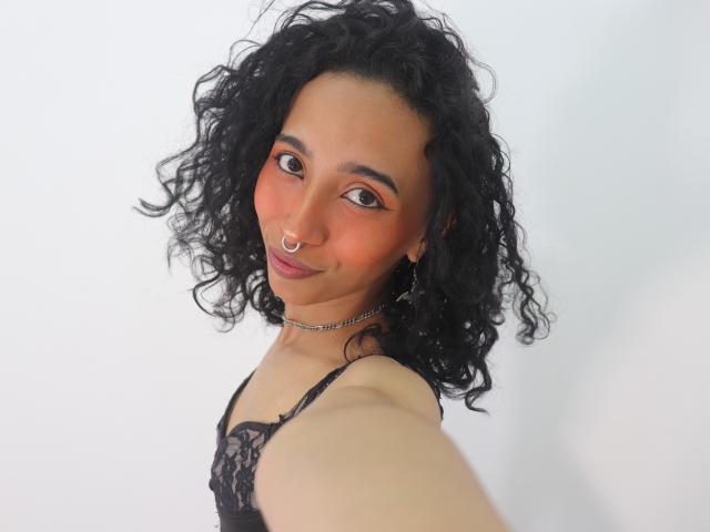 Agnis a les cheveux noirs et bouclés, une peau brune, un anneau au nez et un sourire charmant. Elle porte de la dentelle noire et une chaîne en argent, et regarde l'appareil photo..