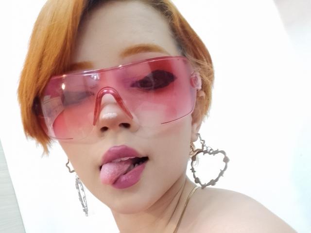 ChloeeWilliams with orange hair, pink transparent visor, and silver heart-shaped earrings, tongue out, pink lipstick, looking up, showing her nose and slightly open mouth.