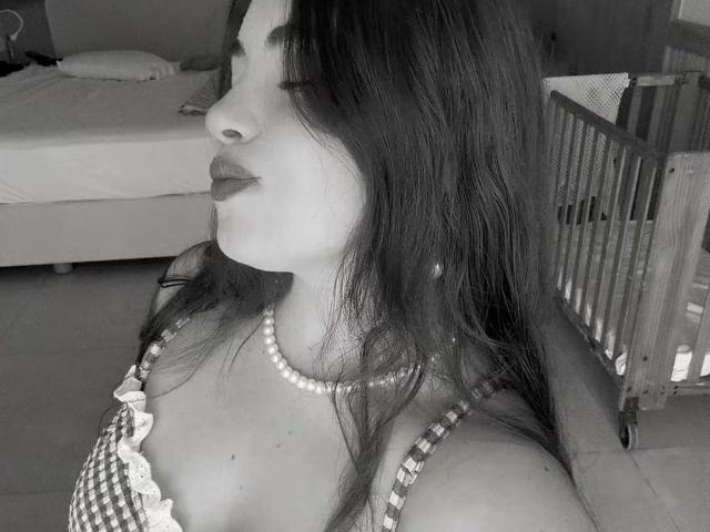 VicluX, avec de longs cheveux noirs, portant un haut à carreaux avec de la dentelle, un collier de perles et du rouge à lèvres foncé, regardant de côté, montrant sa poitrine et son nez sur une photo en noir et blanc..