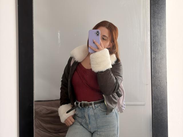 VicluX, avec des cheveux roux, portant une veste marron avec un col en fausse fourrure blanche, un haut rouge et un jean taille haute, prend un selfie devant un miroir avec un téléphone violet. Les seins sont couverts par le haut..