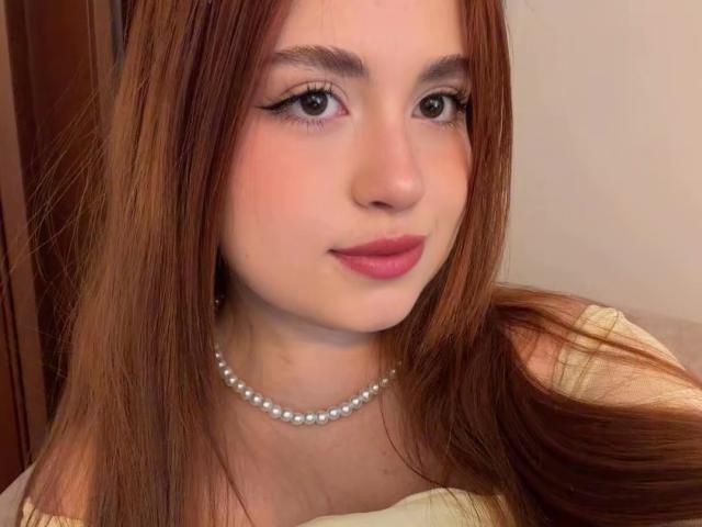 VicluX a les cheveux bruns clairs, les yeux verts et porte du rouge à lèvres rose. Elle porte un collier de perles et un haut de couleur crème, et regarde l'appareil photo avec un léger sourire..