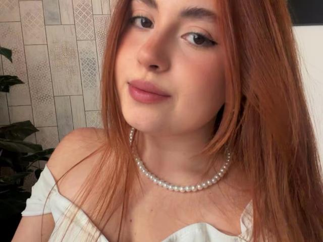 VicluX ha i capelli rossi, la pelle chiara e indossa una collana di perle con una maglietta bianca a spalle scoperte. Ha il rossetto rosa e un leggero sorriso..