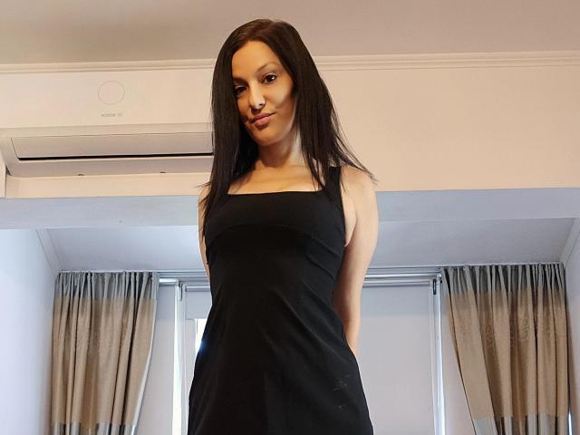 JoliDanielle with long black hair, wearing a black dress, standing with hands in pockets, looking down with a slight smile, her tits and ass are not visible.