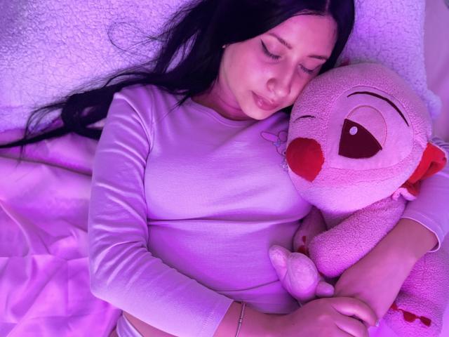 LucyGrey69, con lunghi capelli neri, occhi chiusi, indossa una camicia viola e abbraccia un giocattolo di peluche a forma di uccello rosa con il becco rosso, sdraiata su un cuscino bianco con una texture..