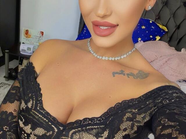 NicolSNaya, con piel morena, labios carnosos y un tatuaje en el hombro derecho, vestida con una blusa negra de encaje con un hombro descubierto, que deja ver sus senos, y un collar de diamantes..