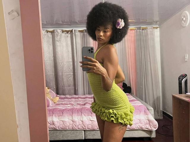 KarlaRicochet, con un afro rizado, vestida con un vestido halter verde con volantes, tomando una selfie en el espejo, mostrando un tatuaje en su muslo, con una flor rosa en el pelo, luciendo adorable..