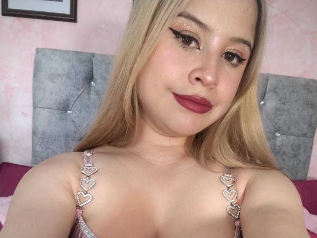 Melani Becker heeft blond haar, roze lippenstift en glinsterende eyeliner. Ze draagt een sprankelend roze top met hartvormige bandjes, waardoor haar kleine borsten zichtbaar zijn. Ze glimlacht lichtjes..