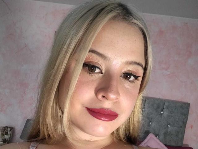 Melani Becker heeft blond haar, bruine ogen en glanzende roze lippenstift. Ze kijkt naar de camera met een lichte glimlach en laat haar schattige, mollige gezicht zien. Haar borsten zijn niet zichtbaar..