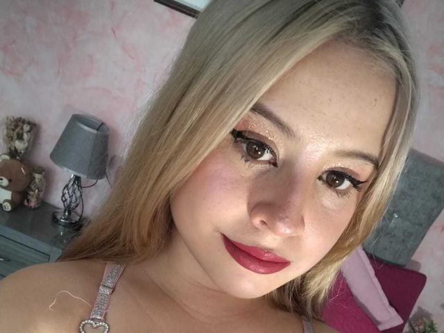 Melani Becker heeft blond haar, bruine ogen en glanzende roze lippenstift. Ze draagt glinsterende oogmake-up en een bandje met een diamantachtige hanger. Haar borsten zijn gedeeltelijk zichtbaar..