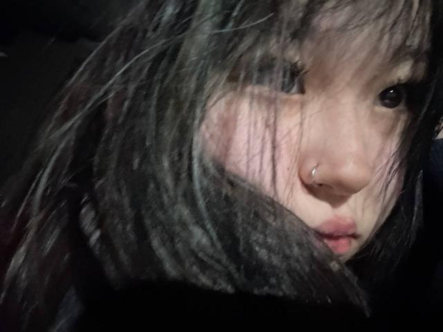 El rostro de JungMcgranor, con cabello negro despeinado, un piercing en la nariz y los labios ligeramente entreabiertos, mirando hacia arriba con ojos oscuros, en un fondo oscuro y borroso. Los senos no son visibles..