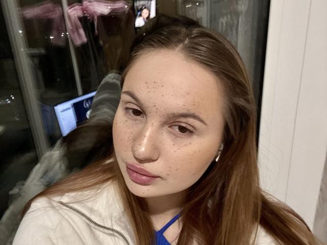 MarisAbnet has brown hair, freckles, and pink lips. She's wearing a white jacket with a blue strap, looking at the camera with a neutral expression.