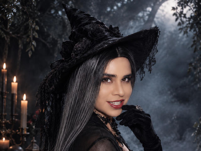 CamilaFetishDirty with long black hair, wearing a black witch hat, black gloves, and a black dress, smiling with red lipstick, candles in background, forest night vibe.