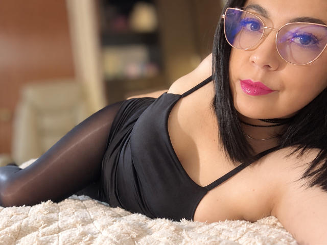BellaFournier, allongée sur un lit, porte un débardeur noir et des collants noirs transparents, avec du rouge à lèvres rose et des lunettes bleues réfléchissantes, un collier noir autour du cou et des cheveux noirs courts..