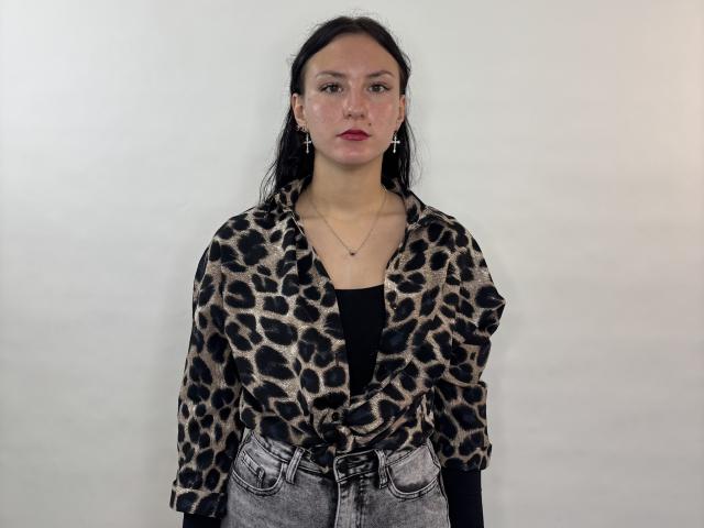 LorisDucking ha i capelli scuri, indossa una camicia a stampa leopardo, una maglia nera e jeans grigi con una cintura, ed è in piedi di fronte a un muro bianco liscio, con un'espressione seria..