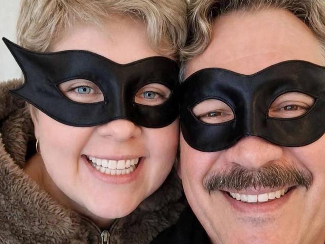 WildLatinCouple ha un viso carino e sorridente, con occhi azzurri, indossa maschere nere, capelli biondi corti e una giacca marrone soffice. WildLatinCouple ha dei baffi e sorride mostrando i denti bianchi..