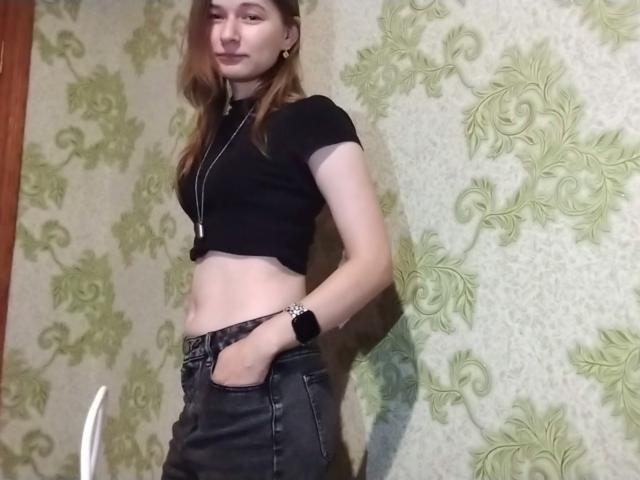 Anna Kleinman, com cabelos castanhos longos, blusa cropped preta que revela seus abdominais planos, vestindo jeans pretos e um colar, em pé com as mãos nos bolsos, olhando para a câmera..