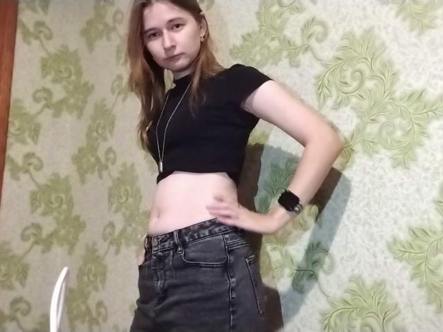 Anna Kleinman, slank, met kleine borsten, draagt een zwarte crop top, waardoor haar buik zichtbaar is, zwarte jeans, handen op haar heupen, bruin haar, een witte ketting, een zwarte horloge, staat, groene behang achtergrond..