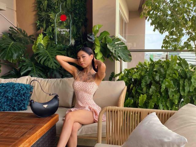 PamelaRougee with tattoos, arms behind head, wearing a pink polka dot dress, sitting on a wicker couch, black handbag on wooden table, green plants, blue pillow, sexy.