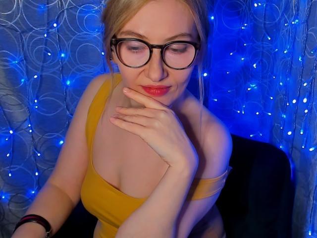 PaulinaSunny with blonde hair, black glasses, yellow top showing her tits, red lipstick, and hand near her face, blue lights in the background. She's sitting on a black chair.