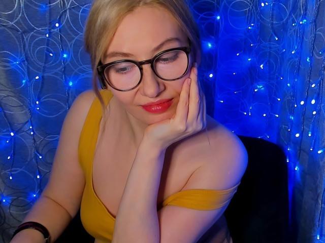Paulina, de cabello rubio, con gafas y labios rojos, vestida con una blusa amarilla, apoyando su mejilla en su mano, sonriendo, con luces de hadas azules en el fondo..