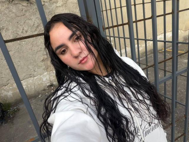 FenixAlthea, avec ses cheveux noirs et mouillés, portant une chemise blanche, regardant l'appareil photo avec un léger sourire, debout près d'une grille métallique, son visage et sa poitrine sont visibles..