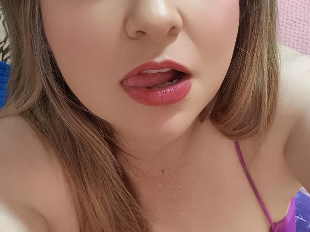 NatalyHami's face, showing pink lips slightly parted, brown eyes, and fair skin, with light brown hair and a pink bra strap visible on her shoulder. Her tongue is peeking out.