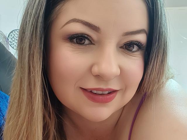 NatalyHami has light brown hair, blue eyes, and pink lipstick, smiling with slightly parted pink lips, wearing a purple top, showing her bare shoulder.