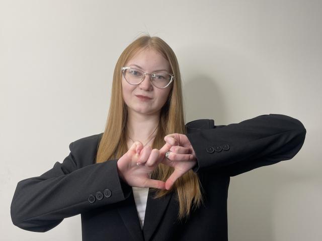 Carmel, mit glattem, blondem Haar, Brille, schwarzem Blazer, weißem Hemd und einem Ring, formt mit ihren Fingern ein Herz, lächelt leicht, hat einen kleinen Hintern und helle Haut..