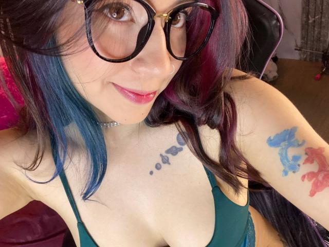 EmmaFeri, com cabelo azul escuro e roxo, óculos, batom rosa, sutiã verde, tatuagens no ombro e no peito, sorrindo, mostrando o colo, sentada em uma cadeira, usando um colar tipo "choker"..