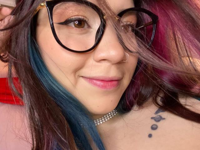 EmmaFeri with black-framed glasses, nose ring, pink lipstick, blue and purple hair, choker, and tattoo on her neck, winking. Boobs not visible.