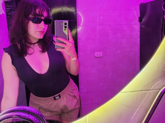 ValeriHairy69, con capelli castano scuro, occhiali da sole neri, un profondo scollo in una maglia nera, pantaloncini rosa, sta scattando un selfie allo specchio in un bagno illuminato di viola. Tiene in mano uno smartphone..
