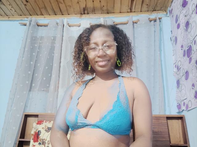 LuciaSexy69 mit lockigem, braunem Haar, Brille, grünen Ohrringen, lächelnd, trägt einen blauen Spitzen-BH, der ihre Brüste zeigt. Sie hat einen mittelbraunen Hautton..