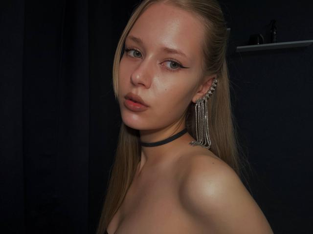 LillianaWhang has pale skin, long blonde hair, and wears a black choker with dangling earrings. She's topless, showing her bare shoulders and small tits. She's looking at the camera.