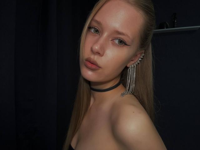 LillianaWhang with blonde hair, black choker, and silver earrings, showing off her bare shoulders and subtle makeup. She's looking at the camera with a slightly parted lip.