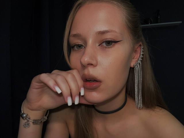 LillianaWhang with light skin, blonde hair, black choker, long eyeliner, white nails, hand near mouth, wearing dangling earrings, and a silver bracelet, looking at camera.