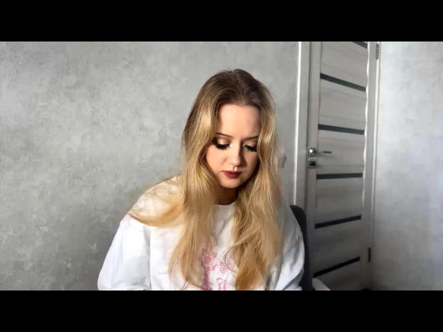 AureliaHavis with long blonde hair, wearing a white shirt, looking down with closed eyes, soft makeup, in a room with a grey textured wall and a white door.