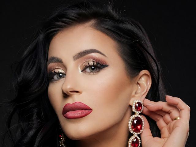 MistressTaylor with dark wavy hair, bold red lipstick, shiny gold eye makeup, and large red gem earrings, touching her ear. Her brown eyes are focused and serious.