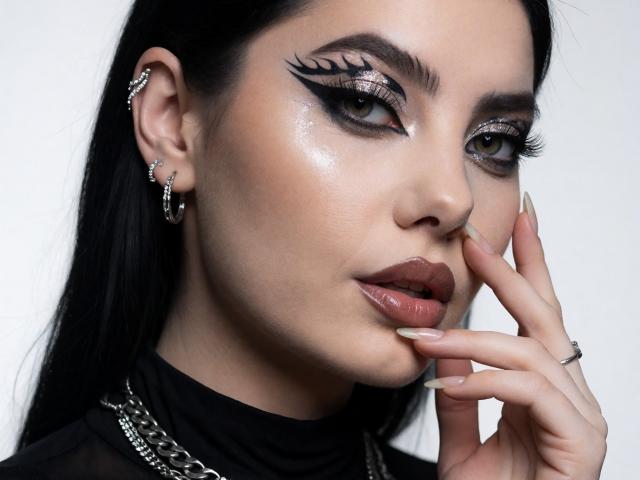 MelissaGlow has dark makeup with dramatic eyeliner, glossy brown lips, and long black hair. She's touching her face with manicured nails, wearing earrings and a chain necklace.