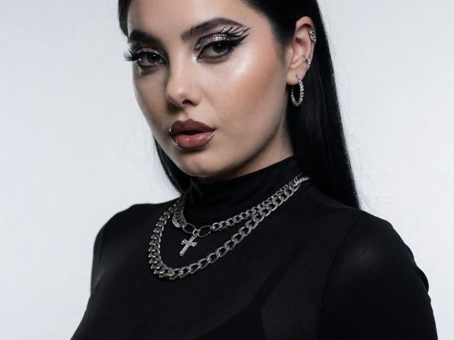 Melissa, met lang zwart haar, donkere eyeliner, glanzende roze lippen, draagt een zwarte doorschijnende top, een zilveren ketting met een kruis en oorringen. Haar gezicht is serieus, ze kijkt vooruit..