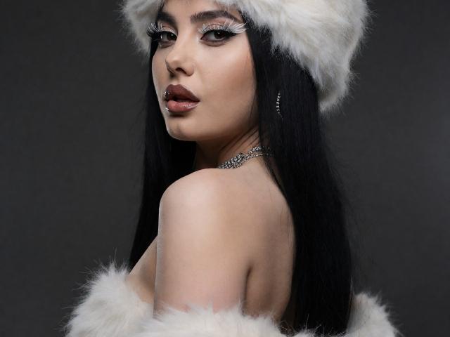 Melissa, mit langem, schwarzem Haar, trägt einen weißen, pelzigen Hut und eine passende, pelzbesetzte Schulterpartie. Sie blickt zurück, mit dunklem Lippenstift und Eyeliner, und zeigt ihre freie Schulter sowie eine glitzernde Halskette..