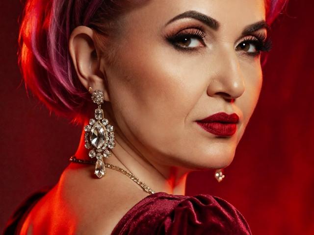 IsabelaConte with purple hair, red lipstick, and diamond earrings, looking over her shoulder. Her tits are slightly visible under a velvet top. Red lighting highlights her face.
