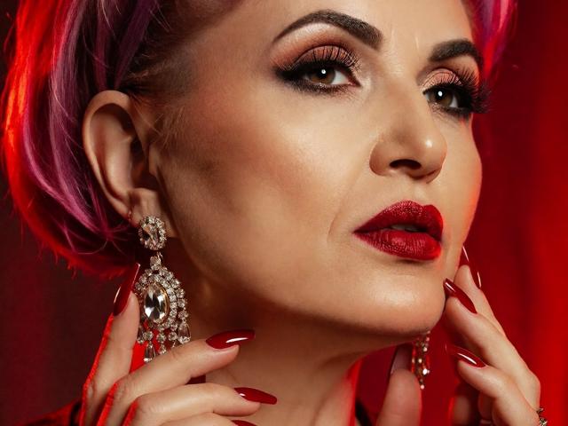IsabelaConte's face with pink hair, red lipstick, and dark eye makeup. She's wearing large, diamond earrings and red nail polish, looking to the side with hands near her face.